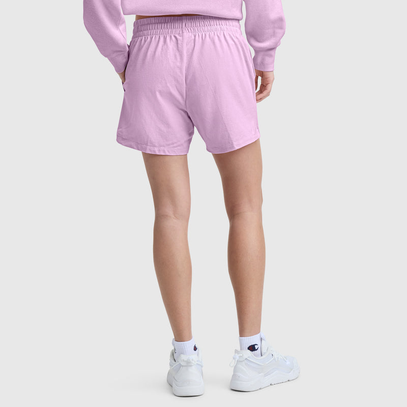 A person wearing Champion 4″ Lightweight Lounge Shorts, C Logo, in light purple along with a matching sweatshirt, white socks, and white sneakers stands on a light gray background with their back to the camera.