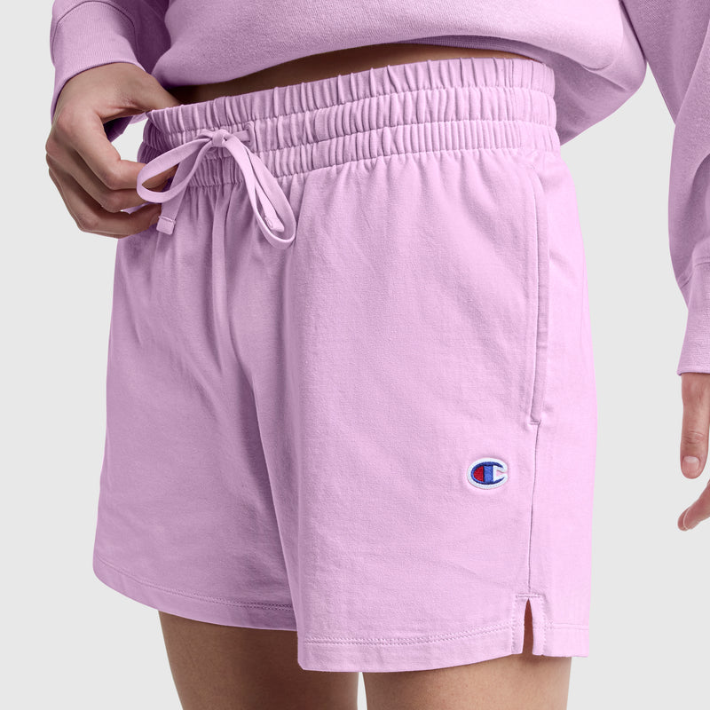 A person is seen wearing Champion’s 4″ Lightweight Lounge Shorts, featuring a light purple hue with an elastic waistband and drawstring. The left leg is embroidered with the iconic C logo, while a coordinating top is partially visible. One hand adjusts the waistband against a plain light gray background. These relaxed women’s shorts from Champion are crafted from lightweight cotton for comfortable everyday wear.