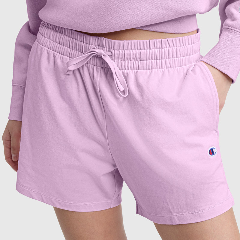 A person is shown wearing Champion’s 4″ Lightweight Lounge Shorts with the C Logo in light purple, featuring an elastic waistband and drawstring, paired with a matching sweatshirt. Only the lower torso and upper legs are visible, with their left hand resting comfortably in the shorts pocket.