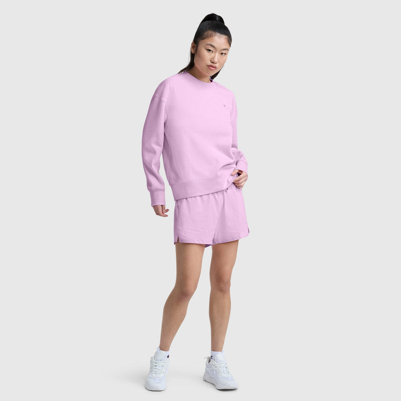 A woman stands against a light gray background wearing Champion’s 4″ Lightweight Lounge Shorts with the C Logo in a light purple shade, paired with a matching sweatshirt and white sneakers. Her hair is styled in a high ponytail, and she is looking at the camera.