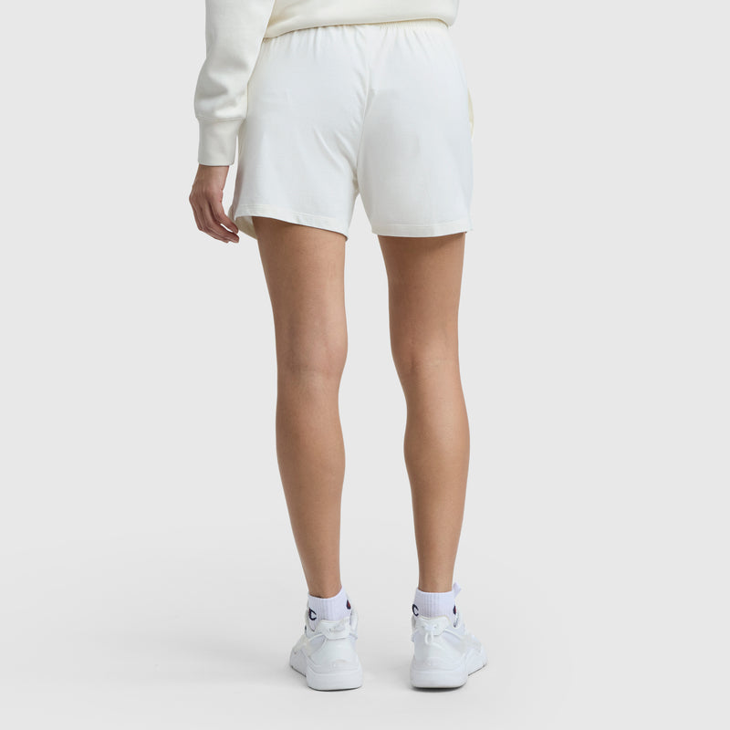 A person stands with their back to the camera, wearing a white sweatshirt, Champion 5" Lightweight Lounge Shorts (C Logo), and white sneakers with socks. The background is plain white.