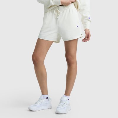 A person, shown from the waist down, wears Champion 5" Lightweight Lounge Shorts with a C Logo, a sweatshirt, crew socks, and sneakers—each featuring small Champion logos—set against a plain white background.