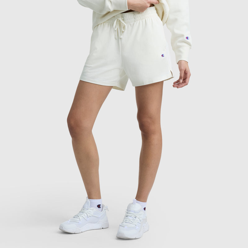 A person, shown from the waist down, wears Champion 5" Lightweight Lounge Shorts with a C Logo, a sweatshirt, crew socks, and sneakers—each featuring small Champion logos—set against a plain white background.