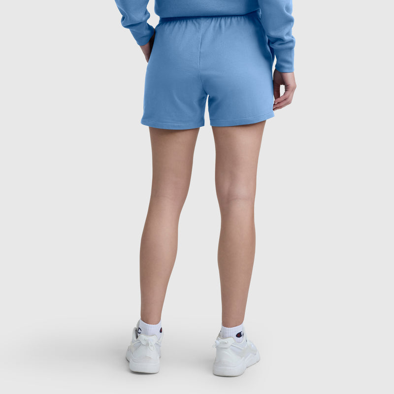 A person stands with their back to the camera, wearing blue Champion 4″ Lightweight Lounge Shorts featuring a comfortable elastic waistband and the signature C Logo. They pair the shorts with a blue sweatshirt, white socks, and white athletic shoes against a plain, light-colored background.