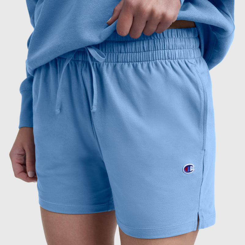 A person wearing Champion’s 4″ Lightweight Lounge Shorts with an elastic waistband and a matching sweatshirt. The light blue shorts feature the C logo on the left leg, and the person’s left hand rests near their pocket.