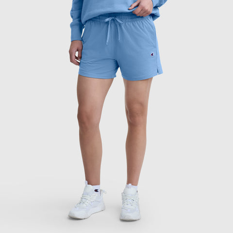 A person wearing a light blue sweatshirt, Champion 4″ Lightweight Lounge Shorts with an elastic waistband, and white sneakers stands against a plain white background.