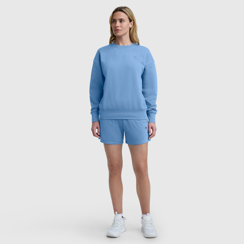 A person stands facing forward against a plain white background, wearing a light blue sweatshirt, Champion 4″ Lightweight Lounge Shorts with the C Logo featuring an elastic waistband, and white sneakers.