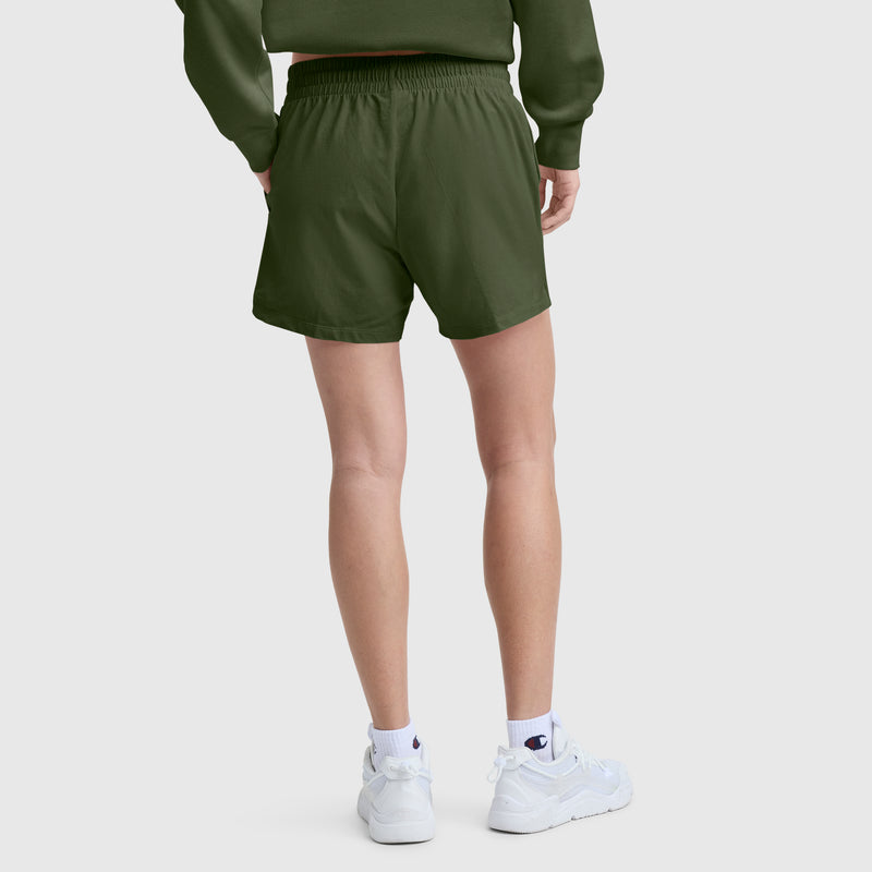 A person stands facing away, wearing Champion's 4" Lightweight Lounge Shorts with C Logo in olive green and a matching sweatshirt, paired with white socks and white sneakers against a plain light background.