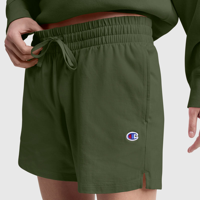 A person wearing Champion 4" Lightweight Lounge Shorts, featuring an olive green color, a drawstring waist, and a small C logo on the left leg, shown from the waist down. These shorts are made from a cotton blend for everyday lightweight comfort.