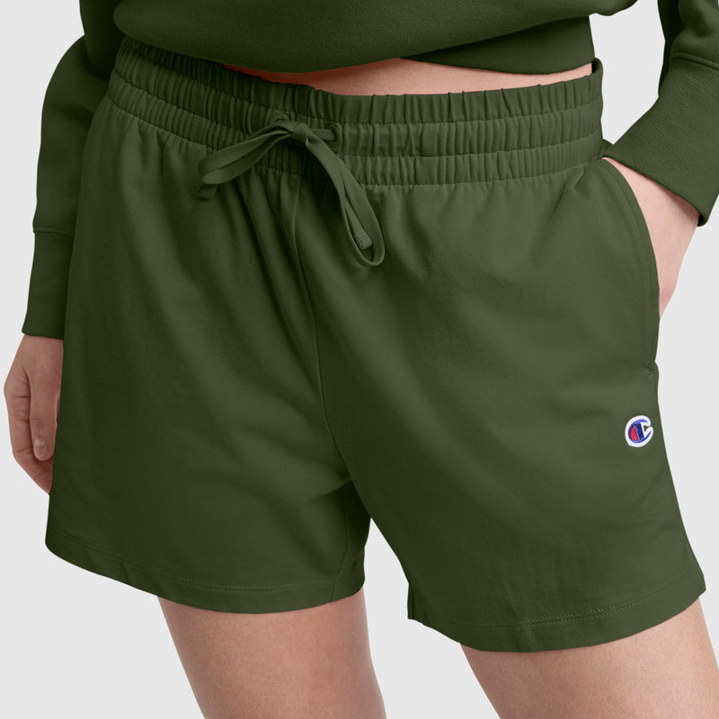 A person wears Champion’s 4" Lightweight Lounge Shorts, C Logo, in olive green—a cotton blend athletic short with an elastic waistband and drawstring, featuring a small Champion logo on the left leg. One hand is tucked into a pocket. The look is completed with a matching green top.