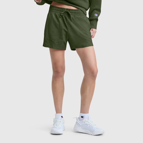 A person stands against a plain light background, showing only their lower body, wearing Champion’s 4" Lightweight Lounge Shorts with C Logo in olive green, paired with a matching green sweatshirt, white Champion sneakers, and white socks.