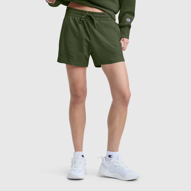 A person stands against a plain light background, showing only their lower body, wearing Champion’s 4" Lightweight Lounge Shorts with C Logo in olive green, paired with a matching green sweatshirt, white Champion sneakers, and white socks.