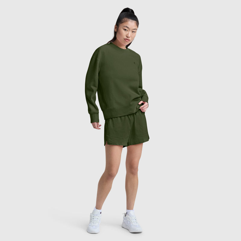 A woman stands against a plain background wearing Champion's 4" Lightweight Lounge Shorts with a matching olive green sweatshirt and white sneakers. Her long dark hair is tied up in a high ponytail.