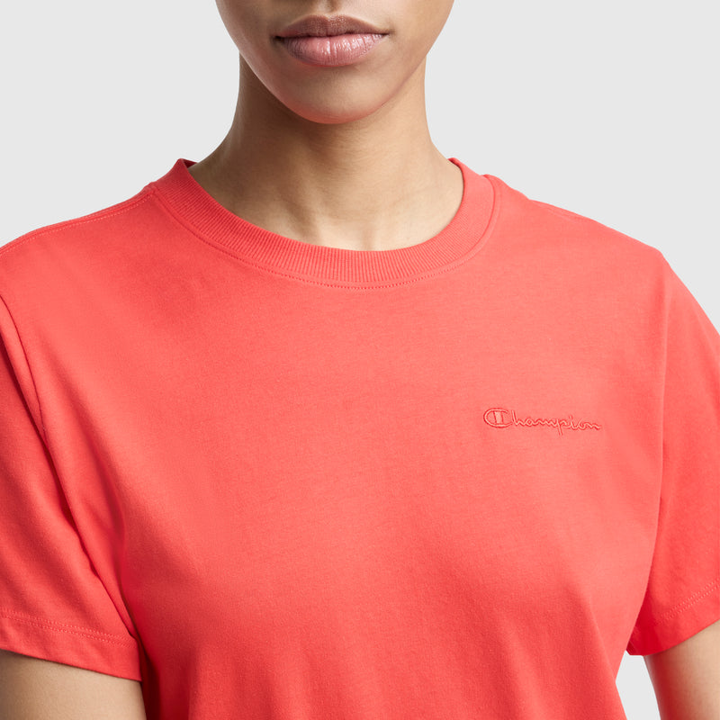 A woman wears the Champion Classic T-Shirt in bright coral pink, shown from the shoulders up against a plain light background. The shirt features a tonal script logo embroidered on the left chest.