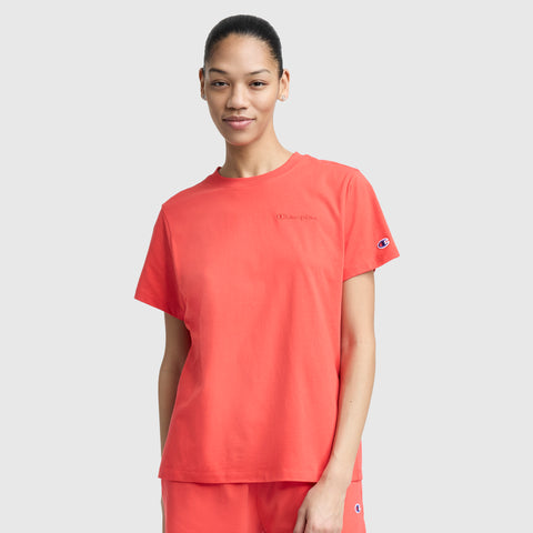 A woman in a loose-fitting coral Champion Classic T-Shirt with tonal left chest script and matching shorts stands against a light background, smiling softly. The t-shirt features subtle brand embroidery on the chest and sleeve.