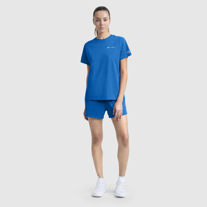 A woman stands against a plain background in a matching bright blue Champion set, featuring the Classic T-Shirt with a tonal left chest script and shorts, made from a soft cotton blend, styled casually with white sneakers.