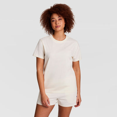 A woman with curly hair wears a Champion Classic T-Shirt with tonal left chest script and matching shorts, standing against a plain white background and looking at the camera with a neutral expression.