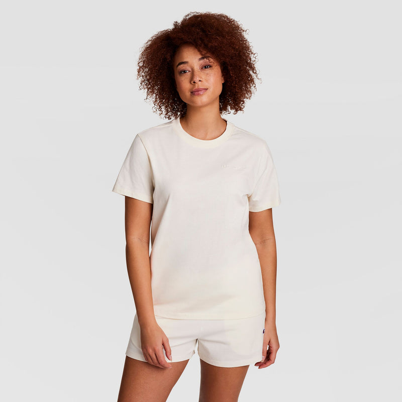 A woman with curly hair wears a Champion Classic T-Shirt with tonal left chest script and matching shorts, standing against a plain white background and looking at the camera with a neutral expression.