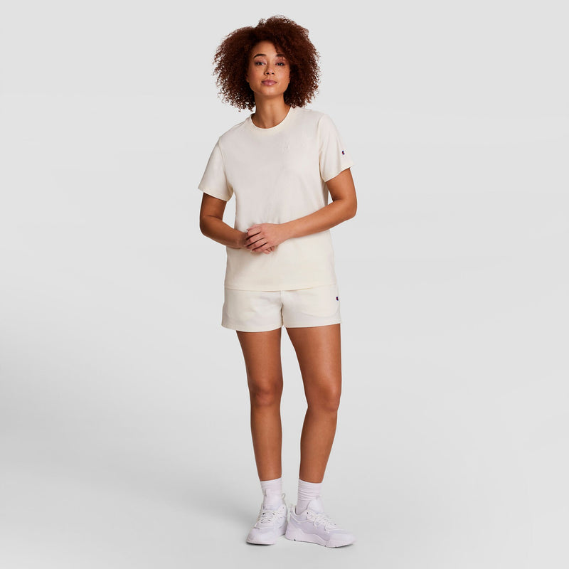 A woman with curly hair stands facing forward, wearing a Champion Classic T-Shirt with tonal left chest script, matching shorts, white crew socks, and white sneakers against a plain light background.