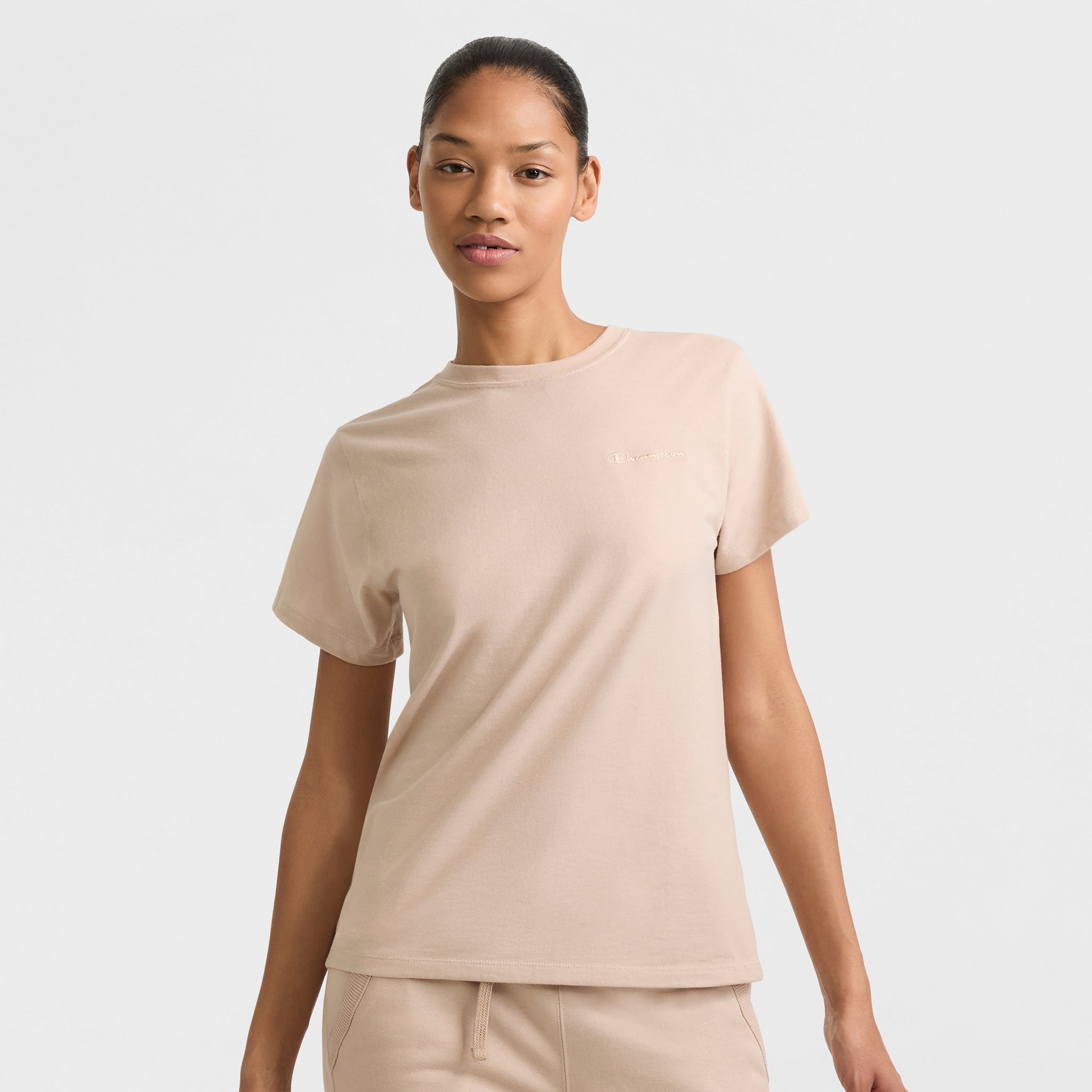 A woman wears the Champion Classic T-Shirt with Tonal Left Chest Script in beige, paired with matching beige pants, standing against a plain light background and looking at the camera with a relaxed expression.