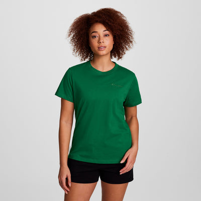 A woman with curly hair wears a green Champion Classic T-Shirt, Tonal Left Chest Script, and black shorts, standing against a plain light gray background.