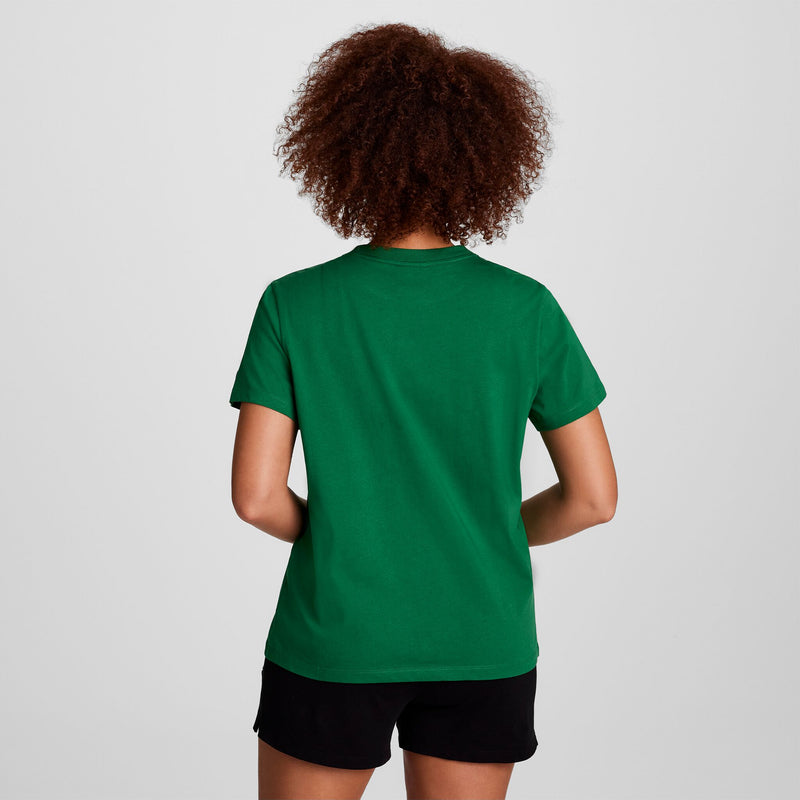 A person with curly hair faces away, wearing a green Champion Classic T-Shirt with tonal left chest script and black shorts, set against a plain light background.