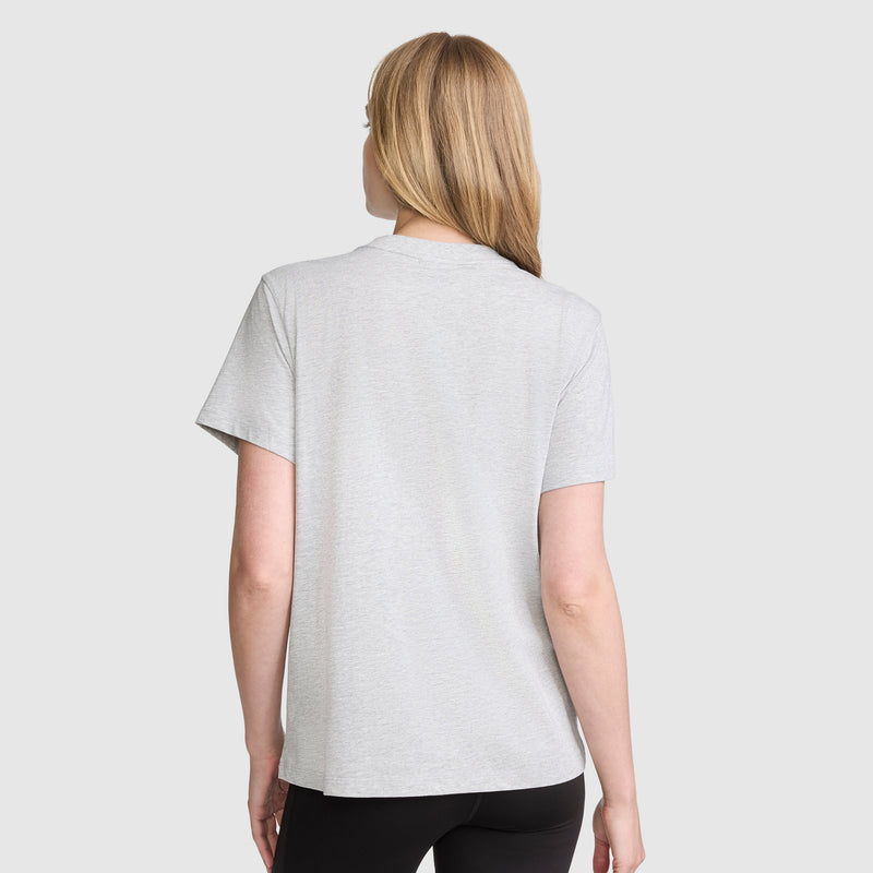 A woman with straight blonde hair is standing with her back to the camera, wearing a Champion Classic T-Shirt, Script Logo in light gray and black pants against a plain light background.