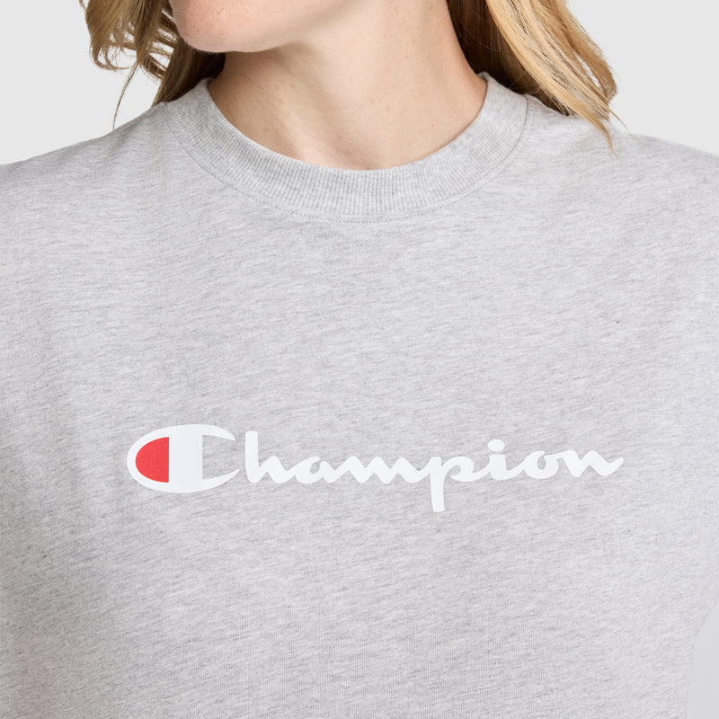 A person wearing a light gray Champion Classic T-Shirt, Script Logo, featuring the white and red script logo across the chest. The person's face is partially out of frame.
