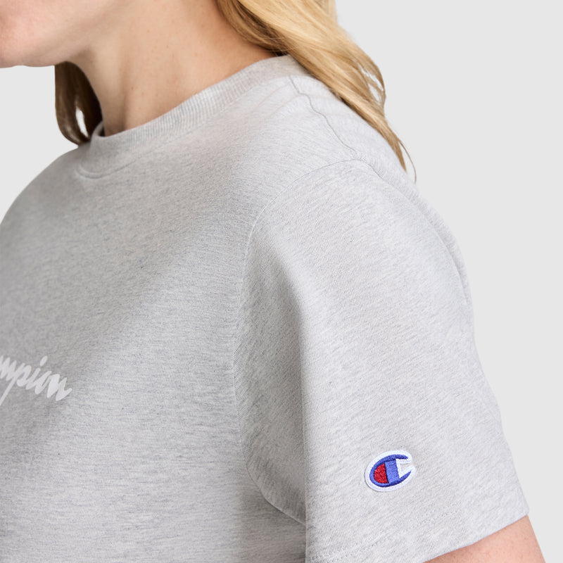 Close-up of a person wearing the Champion Classic T-Shirt, Script Logo in light gray. The iconic script logo patch appears on the sleeve. Only the person's mouth, chin, and hair are visible; their face is not fully shown.