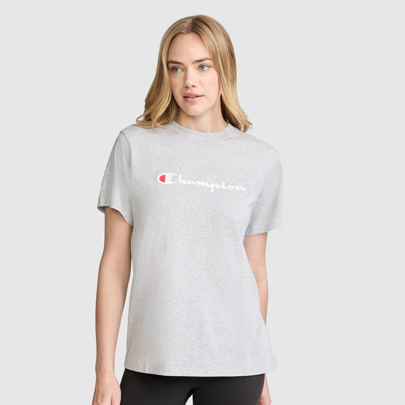 A blonde woman stands against a plain light gray background wearing black pants and the Champion Classic T-Shirt, Script Logo in light gray.