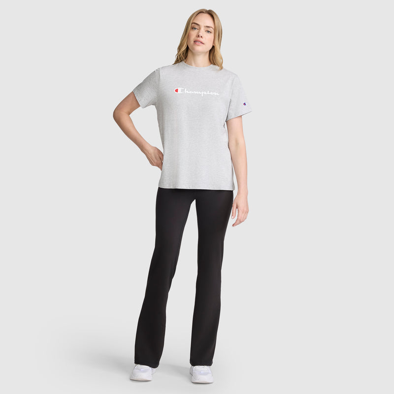 A woman stands facing forward on a plain light background, wearing the Champion Classic T-Shirt with script logo in gray, paired with black pants and white sneakers. Her left hand rests on her hip while her right arm hangs by her side.