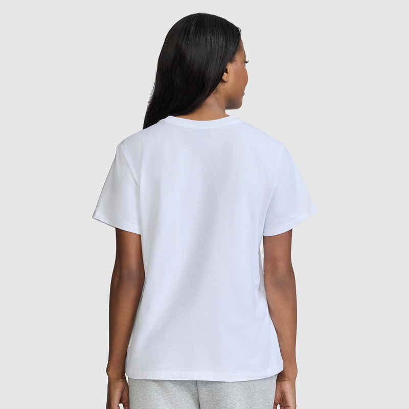A person with long dark hair is standing and facing away, wearing a plain white Champion Classic T-Shirt, Script Logo paired with light gray pants, against a light gray background.