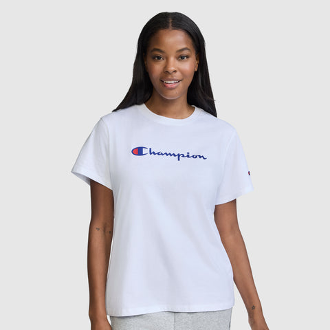 A woman with long dark hair is standing and smiling against a plain light gray background, wearing the Champion Classic T-Shirt, Script Logo, paired with light gray pants.