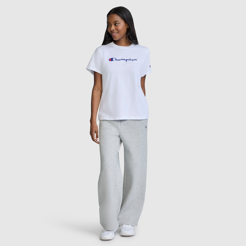 A person stands smiling, wearing the Champion Classic T-Shirt, Script Logo in white, paired with light gray sweatpants and white sneakers, against a plain light gray background.