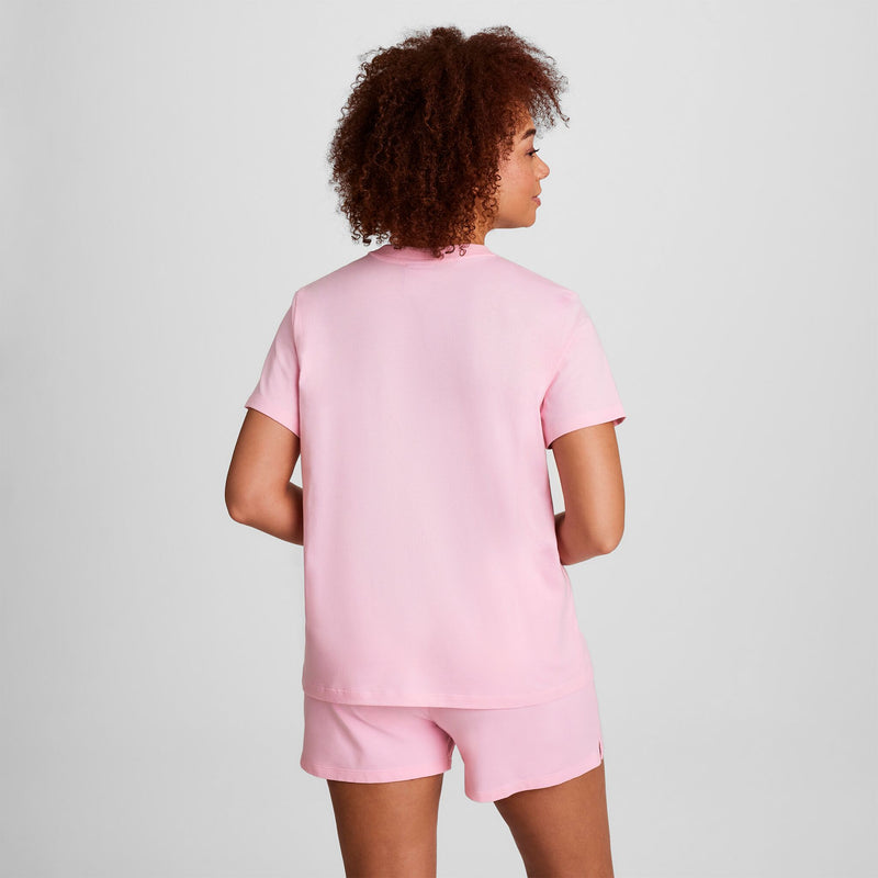 A person with curly hair is standing with their back to the camera, wearing a light pink short-sleeve shirt and matching shorts against a plain light background.