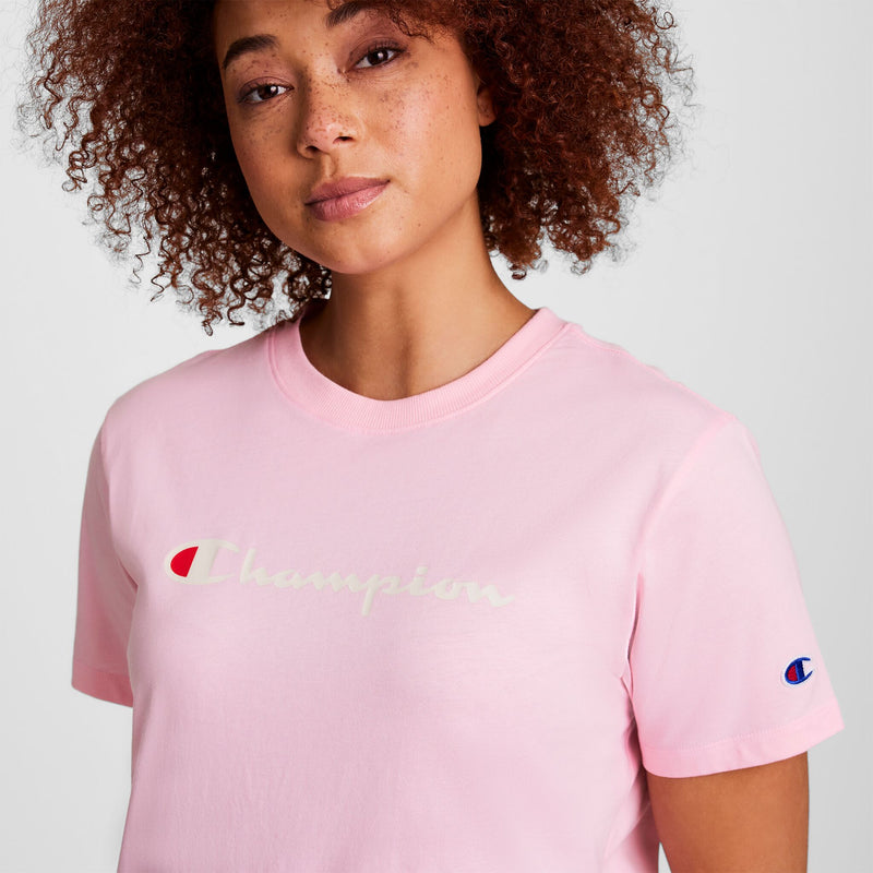 A woman with curly hair wears a light pink Champion t-shirt with the brand’s logo in white across the chest and a small embroidered logo on the sleeve. She looks slightly to the side with a neutral expression.
