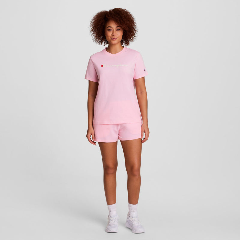 A person with curly hair wearing a light pink T-shirt and matching shorts stands facing forward. They are also wearing white socks and white sneakers against a plain light background.