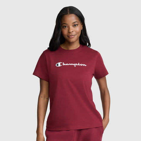 A woman with long dark hair stands against a plain light gray background, smiling slightly while wearing the Champion Classic T-Shirt, Script Logo in maroon along with matching pants.