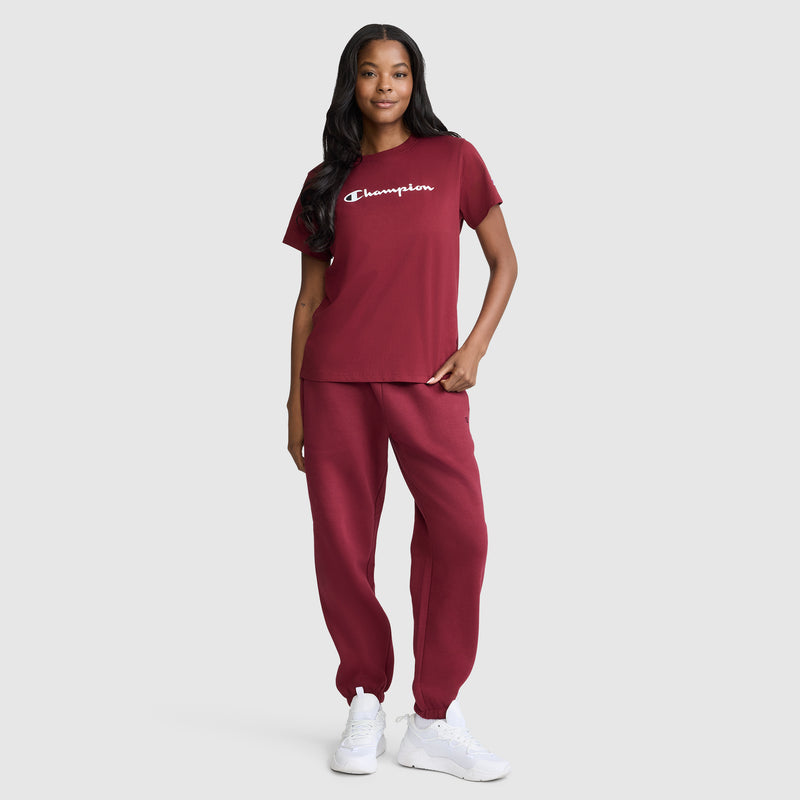 A woman stands and smiles, wearing a matching maroon Champion Classic T-Shirt with the signature script logo and jogger pants, paired with white sneakers, set against a plain light gray background.