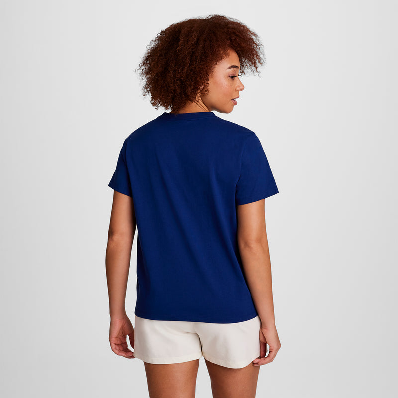 A person with curly hair stands with their back to the camera against a plain white background, wearing a dark blue Champion Classic T-Shirt, Script Logo, paired with light-colored shorts.