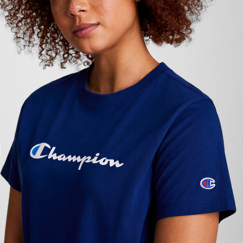 A person with curly hair wears a blue Champion Classic T-Shirt, Script Logo, which features the signature script logo across the chest and a small Champion “C” logo on the sleeve. The background is plain and light-colored.