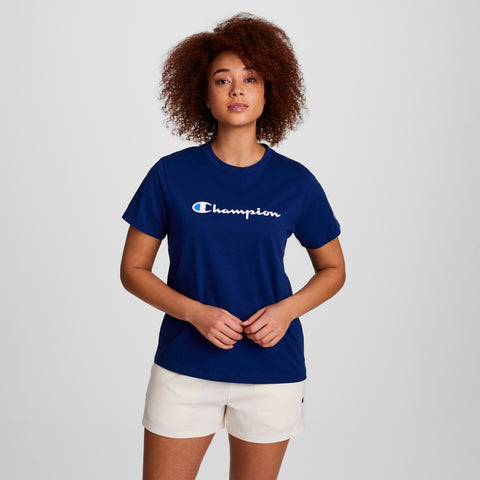A woman with curly hair stands confidently against a plain light background, wearing a blue Champion Classic T-Shirt with the iconic script logo and white shorts, her hands clasped in front as she looks at the camera.