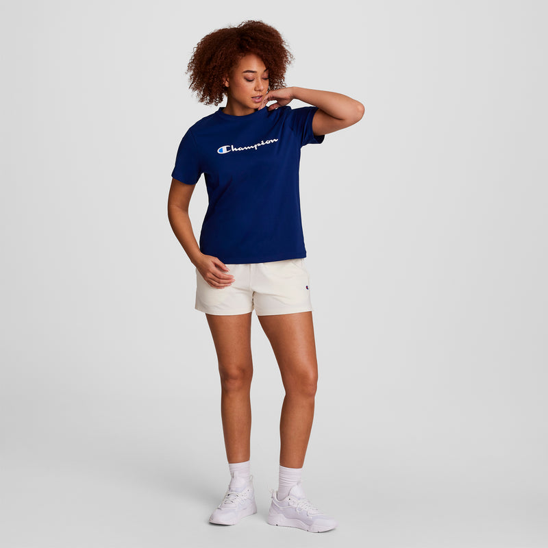 A person stands against a plain background wearing the Champion Classic T-Shirt, Script Logo in blue, paired with cream shorts, white socks, and white sneakers; one hand touches their neck while the other rests by their side.