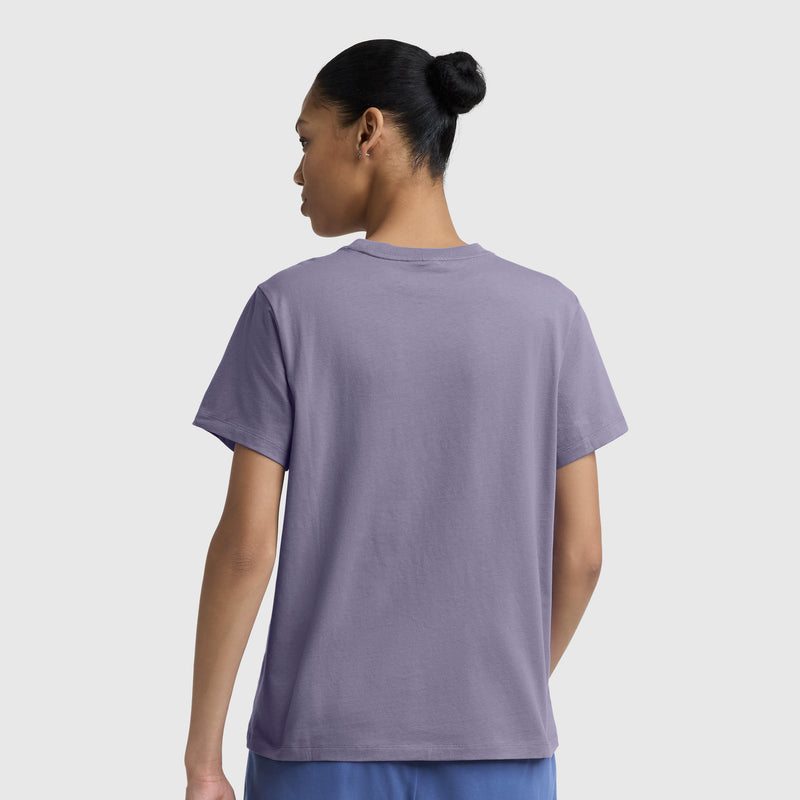 A person with dark hair in a bun is seen from behind, wearing a lavender Champion Classic T-Shirt with script logo and blue pants, standing against a plain light gray background.