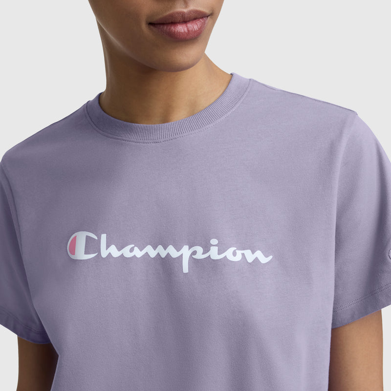A person wears the Champion Classic T-Shirt, Script Logo in light purple, featuring the brand's white script logo across the chest. Only their lower face and upper torso are visible against a plain background.