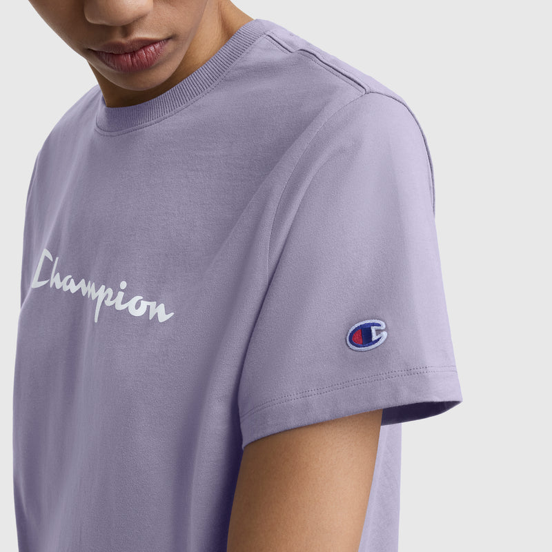 A person wears a light purple Champion Classic T-Shirt, Script Logo—featuring a retro white script logo on the chest and the brand’s sleeve patch—shown from shoulders down.
