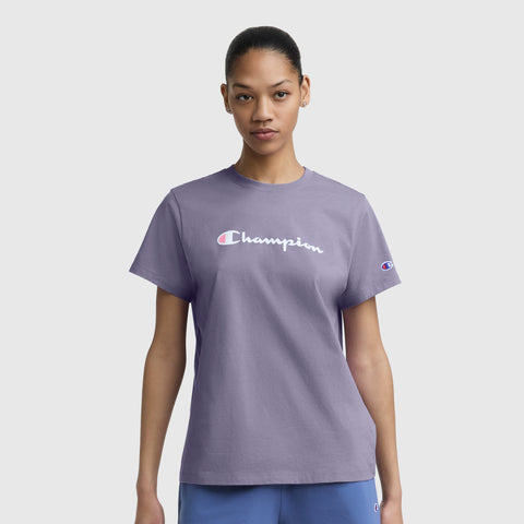 A woman stands against a white background, wearing the Champion Classic T-Shirt with a script logo in lavender. Her hair is tied back as she looks directly at the camera.