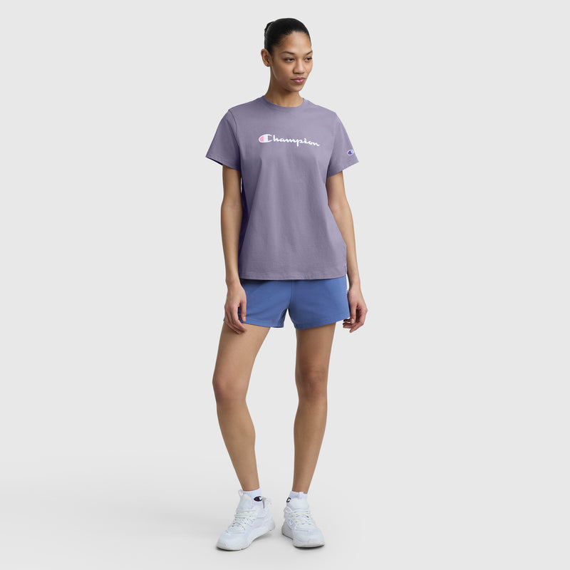 A woman stands against a white background wearing the Champion Classic T-Shirt with Script Logo in purple, paired with blue shorts and white sneakers. Her hair is tied back, and she looks slightly to the side with hands relaxed at her sides.