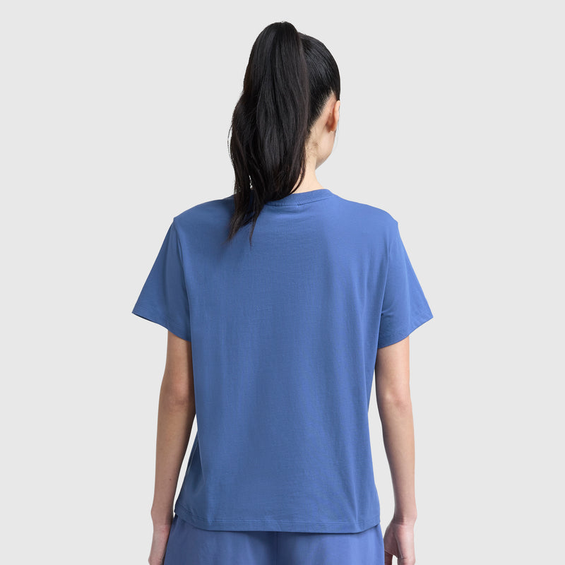 A person with long dark hair in a high ponytail is seen from behind, wearing a Champion Classic T-Shirt with Script Logo and matching pants, set against a plain light background.