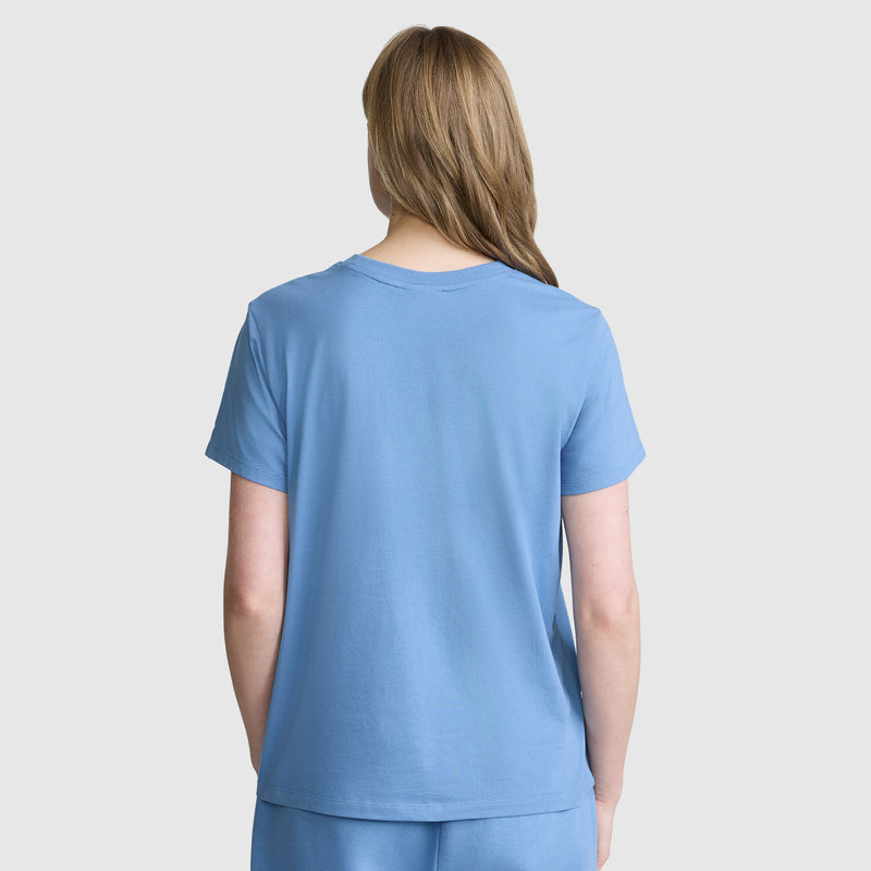A person with light brown hair is seen from the back, wearing the Champion Classic T-Shirt with a subtle script logo in light blue, paired with matching pants, and standing against a plain light background.